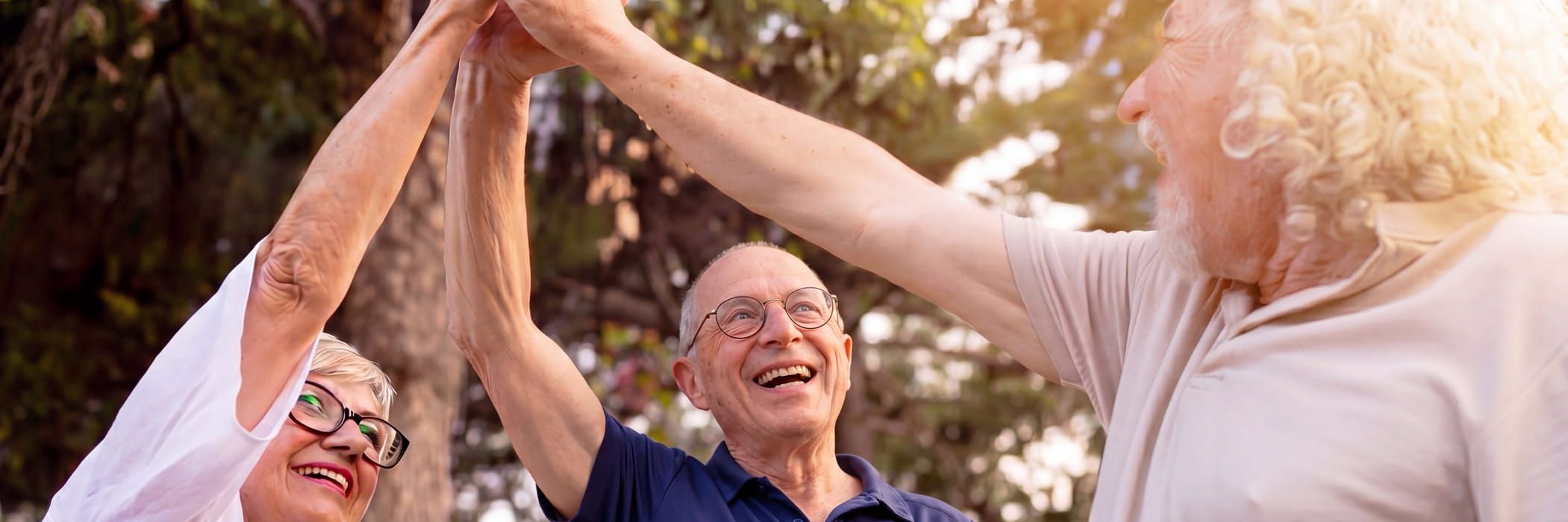 Drie ouderen buiten vormen samen een cirkel met hun armen boven hun hoofd en lachen.